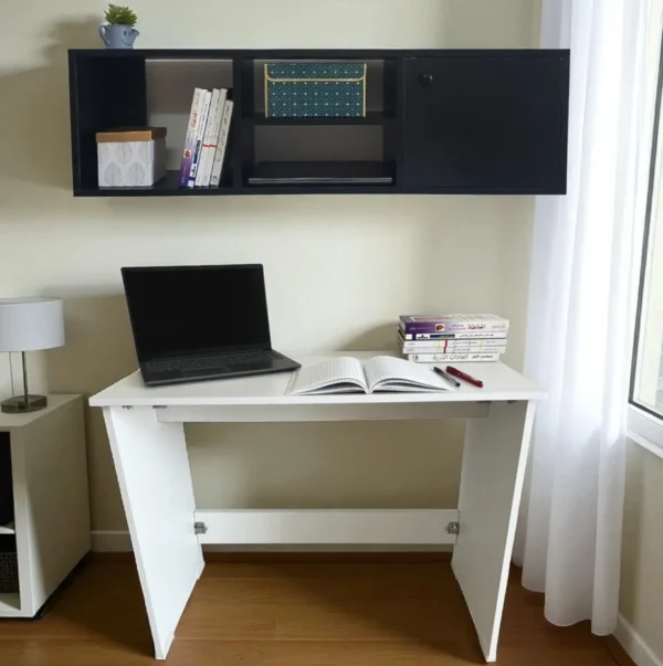Un bureau pliant blanc, avec un ordinateur portable, sous une bibliothèque murale noire avec une porte de placard fermée.