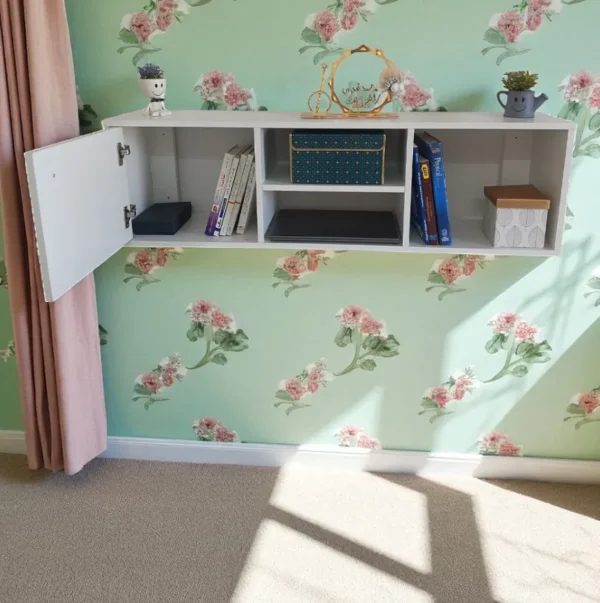 Bibliothèque suspendue blanche sur un mur à motifs floraux verts, avec un placard ouvert et des livres.