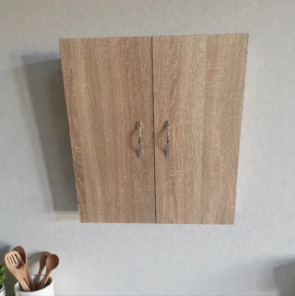 Photo d'une petite armoire de cuisine murale fermée, couleur bois clair chêne, avec deux poignées métalliques, fixée sur un mur gris. Style moderne et épuré.