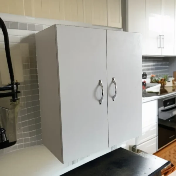 Mini-armoire murale blanche avec poignées chromées, installée au-dessus d'un carrelage gris dans un contexte de cuisine ou de buanderie. Idéal comme meuble pharmacie sécurisé ou pour produits de nettoyage.